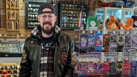 Ashley Hudson Ashley Hudson wearing a cap on backwards behind a stand displaying different trading cards