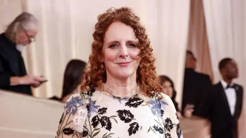EPA Maggie O'Farrell has curled, ginger hair, which is half tied back, and is wearing a white dress with black, white and blue flowers on the pattern. She is smiling at the camera as she stands on a red carpet.