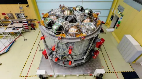 NASA The European Service Module, which is a large cylindrical part of a spacecraft, sits in a cleanroom. On its top surface, several spherical metal tanks are surrounded by metallic insulation. There is shiny silver metallic insulation on its sides too. The module sits on a square platform, and the floor around it is marked with yellow and black safety tape.