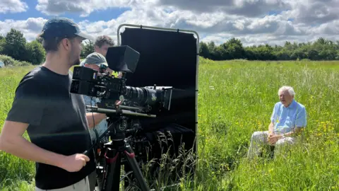 Christian Marot A camera crew are filming Sir David Attenborough sitting in a meadow