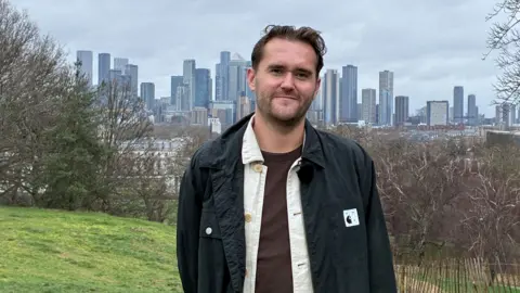 BBC A young white man with brown hair, dressed in brown tshirt, white shirt and black jacket smiles at the camera with a backdrop of trees and skyscrapers