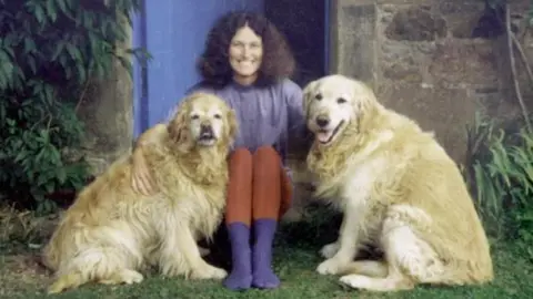 PA Media A woman with long brown curly hair, a lilac top, orange leggings and purple socks is smiling as she sits on the doorstep of a stone-built property which has a light blue-painted door. She has her arm around one of two dogs which appear to be Golden Retrievers. The dogs are sitting to either side of her.