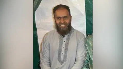 Humayra Ali Man smiles, Masrob, short dark hair and bear. wears grey/pale blue outfit.