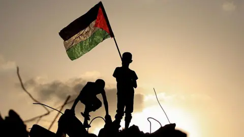 Getty Images Palestinian children play as one waves a Palestinian national flag as he stands on the rubble of a destroyed building at the Bureij camp in Gaza. They are silhouetted against a setting sun illuminates the background of the image.