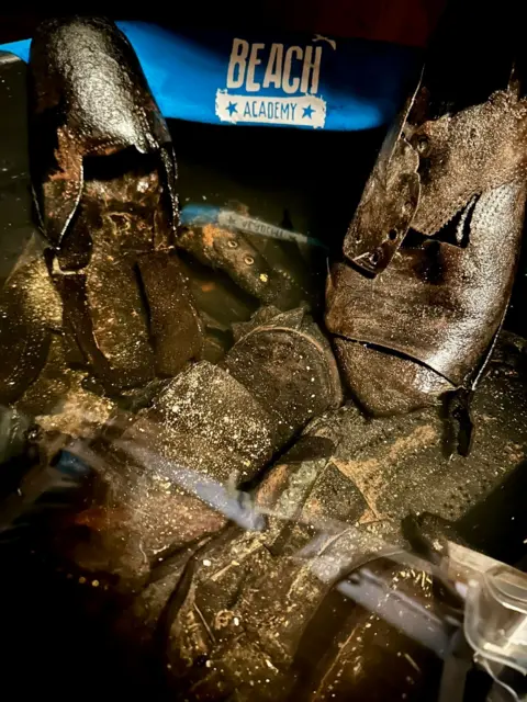 Emma Lamport A black bucket filled with water, with black leather Victorian shoes floating in it and a sign behind which reads Beach Academy