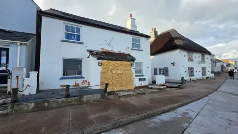 Damaged properties on a seafront. Paint has come away from the front of one property. The front door to the entrance has been boarded up.
