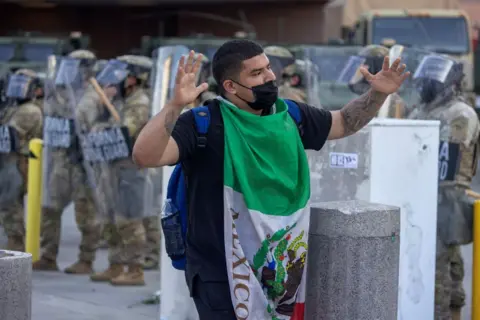 Getty Images A man draped in a Mexico flag with his arms raised walks in front of California National Guard soldiers with shields as protests continue in an approximately one-square mile area of downtown Los Angeles in June 2025