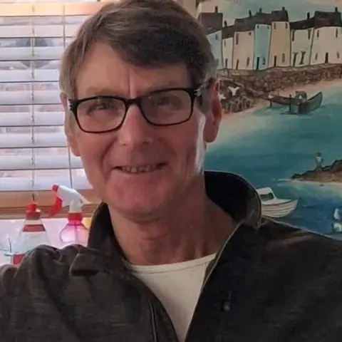 Family Handout A man with brown hair, dark rimmed glasses and a dark jumper smiles into the camera. He is positioned in front of a window with a colourful picture depicting a seaside harbour visible over his shoulder.