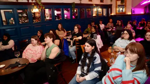 Jonathan Phang A crowd shot of the pub, which shows several tables with people sat around them, watching a TV screen out of shot. The crowd is mostly women, and most are young.