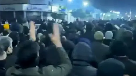 Eyewitness image A large group of people gather in front of buildings in Babol. Someone near the camera has their hands in the air. It is after dark and lights from buildings illuminater the area.