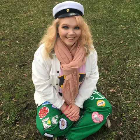 Meri-Tuuli Auer Meri-Tuuli sitting on the grass wearing student party attire consisting of green overalls, a white jacket and a white cap.