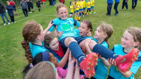 Stephen Tierney Freya is being held aloft by her team mates. She is laughing and has tied back hair. She is wearing a light blue football shirt and bright pink football boots.