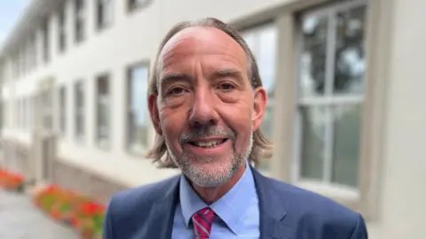 Deputy Steve Falla - A man with long brown hair, with a grey stubbly beard, wearing a blue suit, blue shirt and a red and blue striped tie.