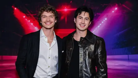 Getty Images Two young men, one in a black suit with open-necked white shirt, and one in a black leather jacket, pose for a photo in front of a large photo of a moodily lit, empty ice hockey rink.