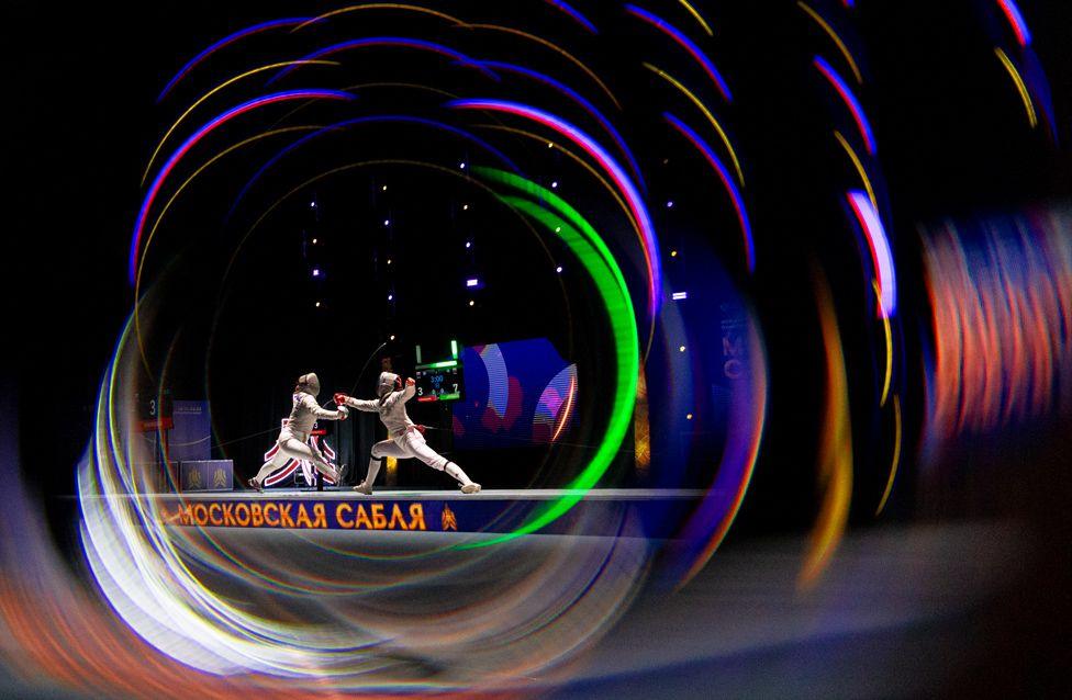 Two fencers in full gear lunge toward each other on a lit piste, framed by swirling colorful light rings.