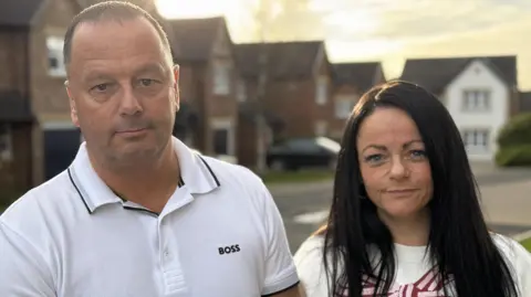 BBC Mike Trotter and Kelly Trotter are standing side by side in a housing estate. Houses and a small road are out of focus behind them. MIke Trotter is 48 and has short cropped hair and is clean shaven and is wearing a white top. Kelly Trotter is 42 and has long dark hair and is wearing a white top with a pink and maroon bow on it.