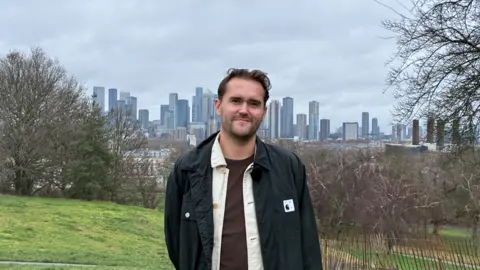 BBC A young white man with brown hair, dressed in brown tshirt, white shirt and black jacket smiles at the camera with a backdrop of trees and skyscrapers
