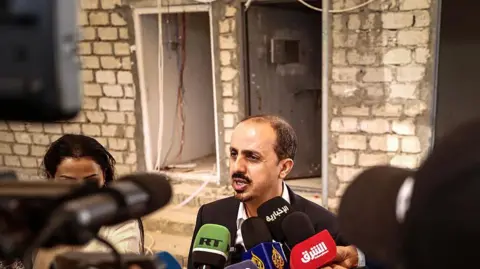 Fadel SENNA / AFP via Getty Image Yemeni Information Minister Moammar al-Eryani speaking with a lot of journalists' microphones in front of him. He is bald with a beard and mustache, and is standing in front of a brick and cement building.