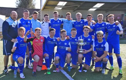 Queen of the South A group of young footballers celebrate winning a trophy. They are in blue kits apart from the goalkeeper who is in pink.
