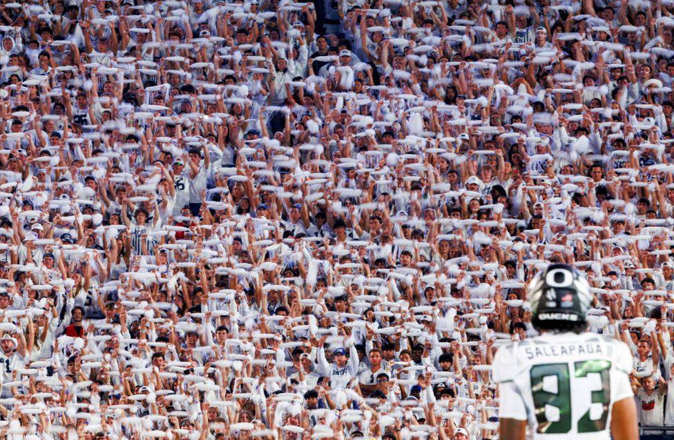 American footballer player Roger Saleapaga stands on the field facing a Penn State University's infamous 'white out' crowd of fans dressed all in white, waving white towels in unison. The player’s jersey displays the number 83.
