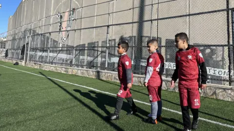BBC News Three Palestinian boys around 10 years old, wearing red football kit, wait in line to take a penalty. They are standing on a green astro turf football pitch with a metal fence behind them. Behind that stands a tall concrete wall