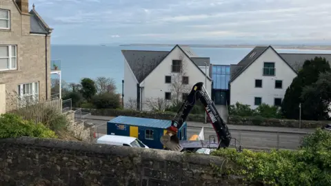 The view from the garden with Godrevy lighthouse in the distance across the sea. In the foreground beyond a boundary wall is a digger at work, a site office container and a white van. Across the road is a large building with an atrium between its two halves.