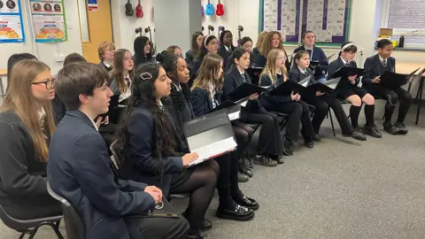 Kate McGough / BBC Around 20 students in school uniform of secondary school age are seated in two rows, facing to the right. They are holding singing sheet folders and mid-song.