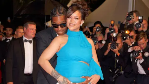 Getty Images A$AP Rocky, wearing a black suits, poses with his partner Rihanna, who is dressed in a bright blue dress. He is hugging her from behind, holding her baby bump.