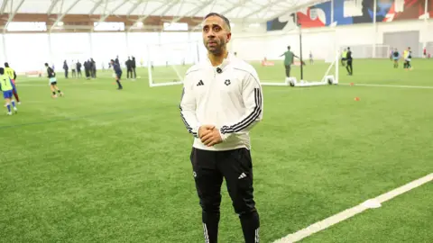 Professional Footballers' Association Riz Rehman wears a white sports jacket with black stripes and is stood on indoor astro-turf. There are players taking shots at a football in the background towards a goalpost