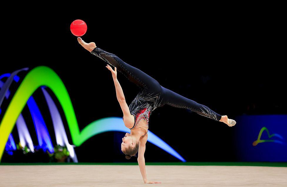 Gymnast Meital Maayan Sumkin performs a one-handed handstand on a mat while balancing a red ball on an extended leg. The gymnast wears a sparkling dark leotard with red accents.