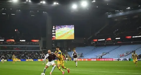 Getty Images An Aston Villa player is tackled by his Maccabi-Tel Aviv opponent. In the background there are other players running towards them, along with Villa Park stands where the crowd can be seen.
