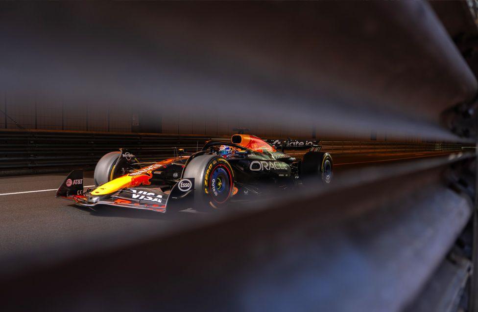 A Red Bull Formula 1 car speeds through a tunnel, captured through the slats of a metal barrier. The car features vibrant red, yellow, and black colors with visible sponsor logos.