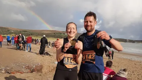Caroline Stafford Gareth and Caroline in running clothes on Swansea beach. They are smiling and holding running medals.