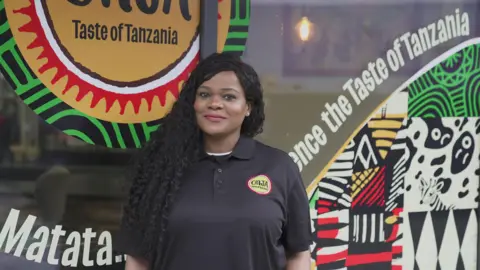 Photograph of Justina John stood outside of her store, Onja. She has long black curled hair, brushed around the side, on her left shoulder. She has brown eyes and smiles at the camera. The restaurant window reads: "Taste of Tanzania" alongside vibrant tribal prints.