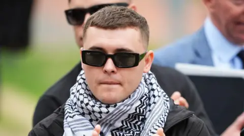 PA Media Liam &Oacute;g &Oacute; hAnnaidh has short dark hair, he is wearing black sunglasses, a black and white scarf and a black coat.