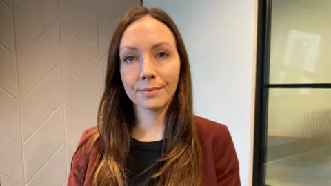 Amy Clowrey, who has long brown hair and wears a red jacket over a black top, looks at the camera.