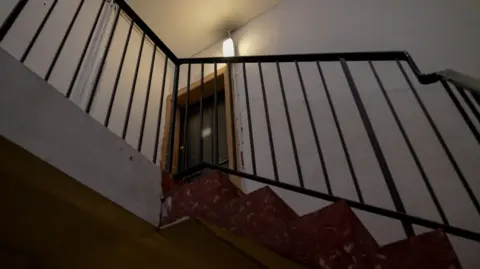 A photo of a flat door on a stairwell landing, taken from below, visible through black handrails. The black door and its yellow frame are chipped and peeling and the walls are a dull white. There is a light above the door.