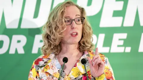 PA Media Lorna Slater who has blonde hair and is wearing black glasses, speaks with a raised pointed finger at a press conference with a green background