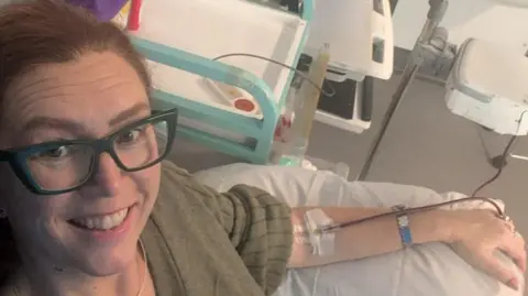 Sian Chathyoka Close up selfie of Sian smiling whilst laying in a hospital bed with a drip going into her left arm. She wears a green knitted jumper and has blue square glasses.