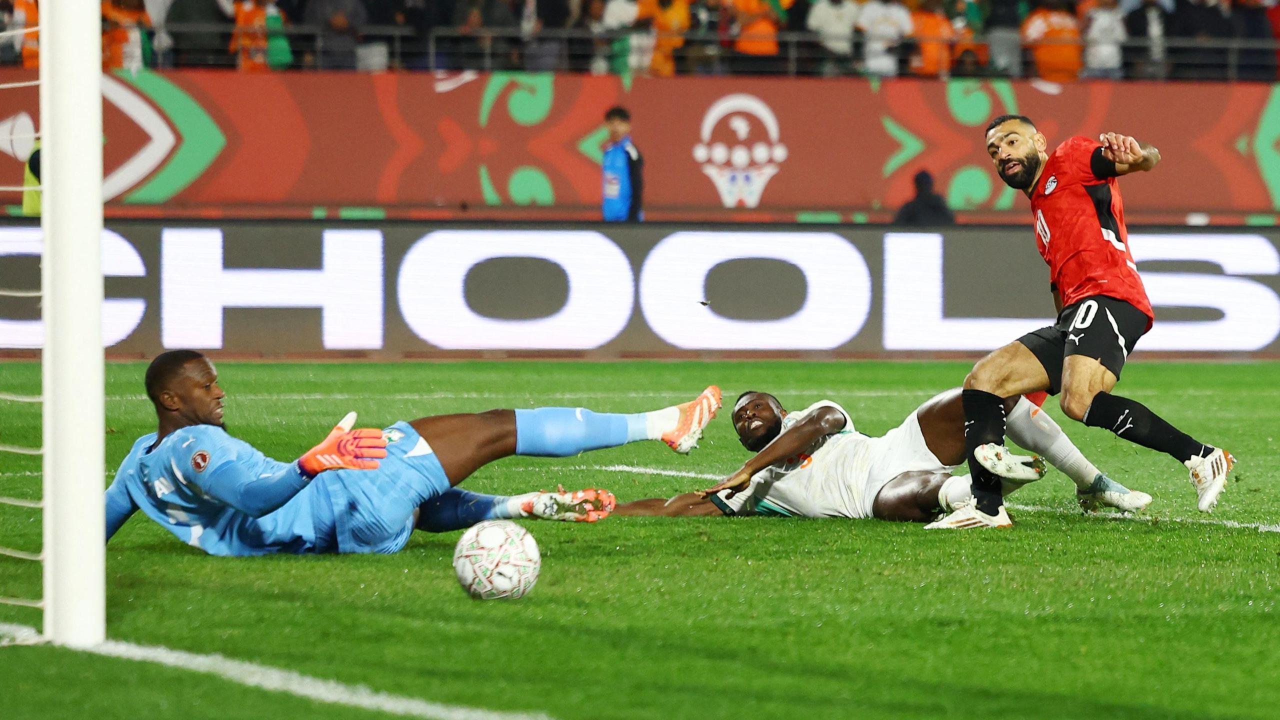 Mohamed Salah scores a goal for Egypt against Ivory Coast at Afcon 2025. Salah, wearing a red Egypt jersey with black short and socks and white boots, is on the far right of the image, falling over slightly to his left while watching the ball travelling towards goal. An Ivorian defender in all-white is sliding in along the ground in a failed attempt to block the shot while goalkeeper Yahia Fofana is also on the green turf, dressed all in sky blue. The ball is travelling across the turf, past all three players, heading towards the goal which can only be partially seen on the far left of the image