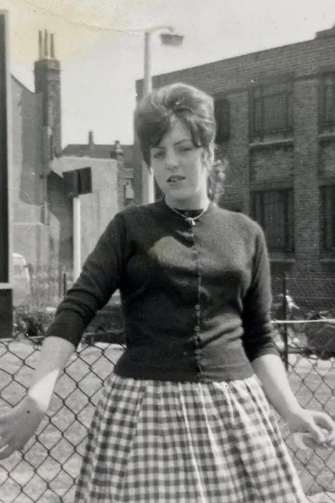 Theresa Fazzani/SWNS A black‑and‑white photograph of a woman standing outside in front of a fence. She has her hair styled in an up do and is wearing a fitted cardigan and a full, gingham‑patterned skirt. Behind her are brick buildings, chimneys, and street signs.