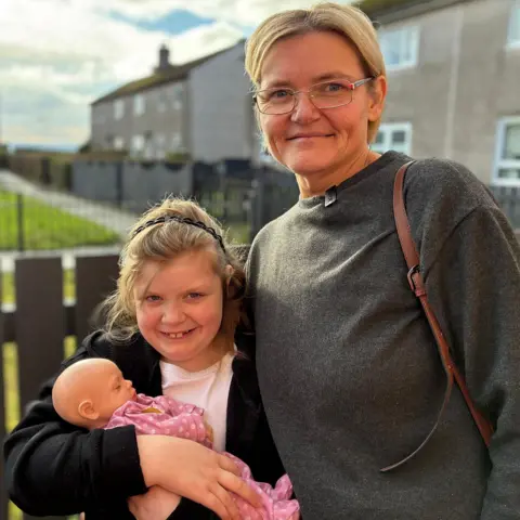 A young girl holding a doll, standing next to a woman with blonde hair and glasses