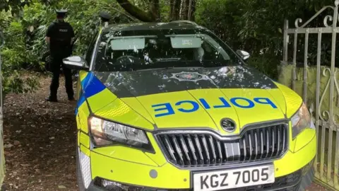 It shows a yellow and blue police car with a uniformed male officer standing with his back to it in the background in front of trees. An open garden gate can be seen to the right of the picture.
