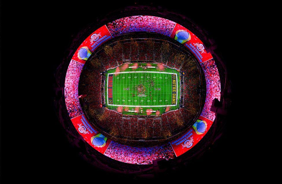 A fisheye view of a brightly lit football stadium packed with fans. The green field is at the center, surrounded by vibrant red and blue banners displaying team names and championship graphics.
