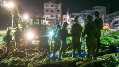 Israel Defense Forces Israeli soldiers standing near an excavator in open ground at night-time, during the search for the body of missing hostage Ran Gvili
