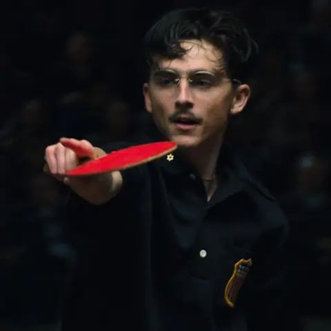 A24 A still from Marty Supreme showing Timothee Chalamet's character playing table tennis, pointing with a table tennis bat in one hand