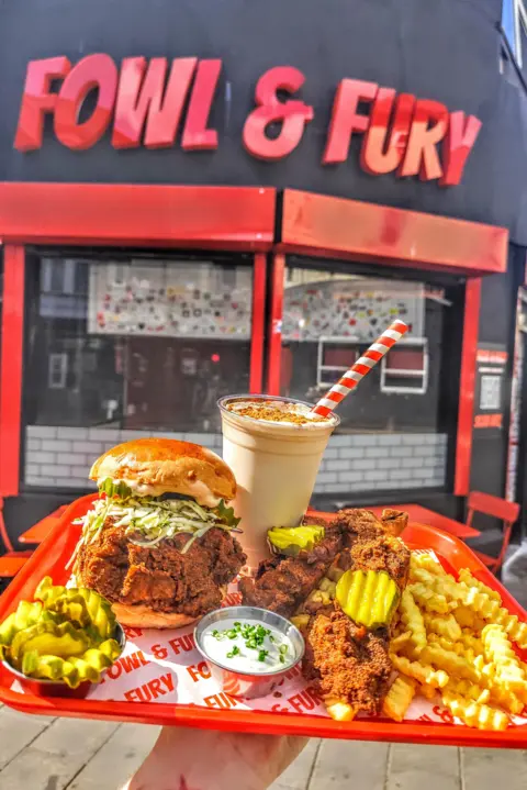Fowl and Fury Fowl and Fury sign, red letters on a black building. Someone is holding up a tray stacked with a chicken burger, chips, gherkins and a milkshake.