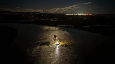 EDF Drone style photo of a boat with the huge cylinder reactor on it. It is dark and there's a bright light shining on the reactor and around it. On the horizon bright lights can be seen on the construction site, including lit-up cranes.