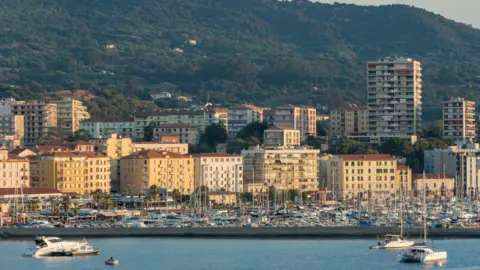Delmarty via Getty Images The evening sun shines on yellow blocks of flats on the coast of Ajaccio, with the marina and boats in front