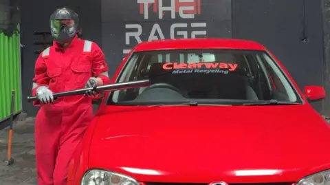 Shuka Piryaee Picture of a woman in a helmet and protective gear that's red. She is holding a baseball bat next to a car.